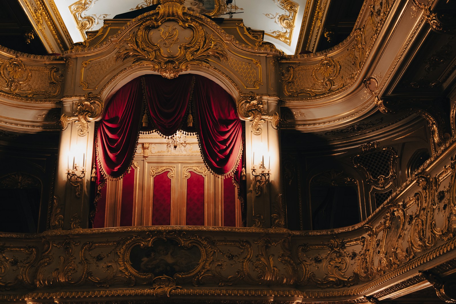 The interior of an ornate opera house with gilded balconies, red velvet seats, an orchestra pit visible in the foreground and the heavy stage curtain partially raised under warm theatrical lighting.